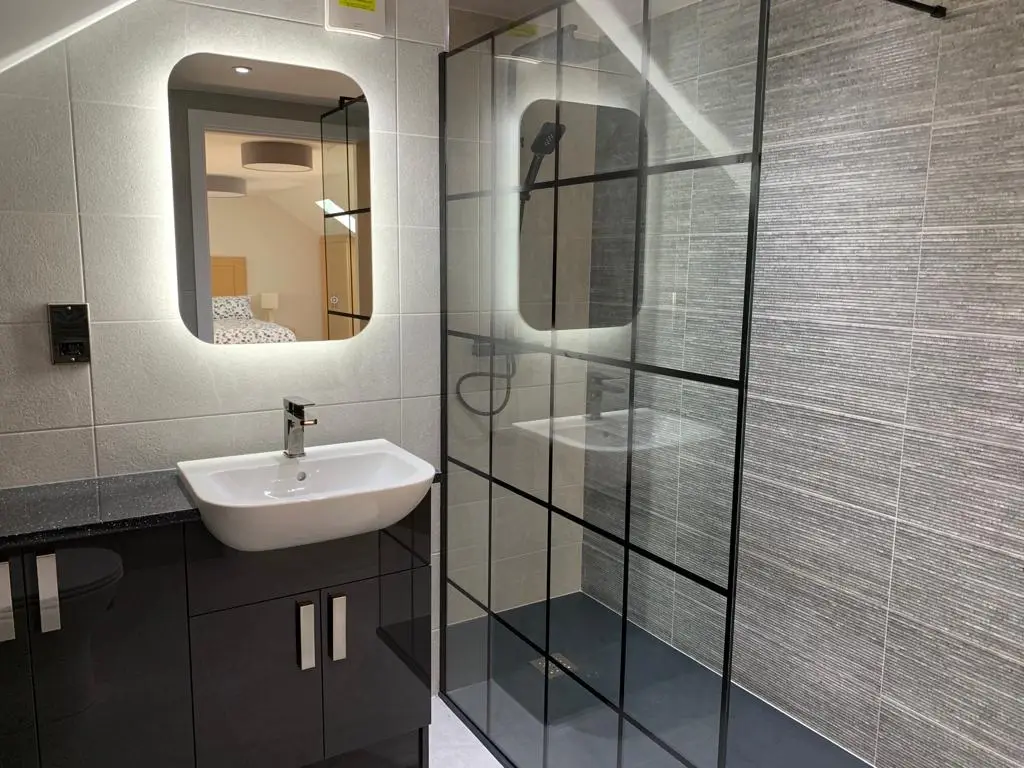Crittall-style black-framed shower screen beside a backlit mirror reflecting the loft bedroom